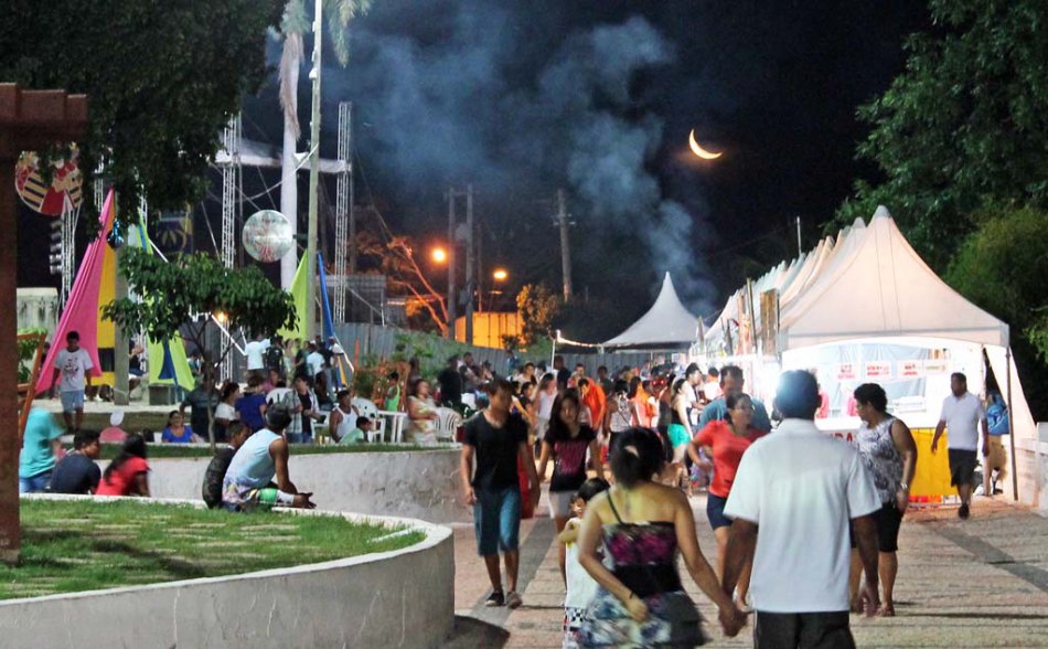 Público caminhando à noite entre tendas brancas e uma praça iluminada, com a lua crescente no céu.