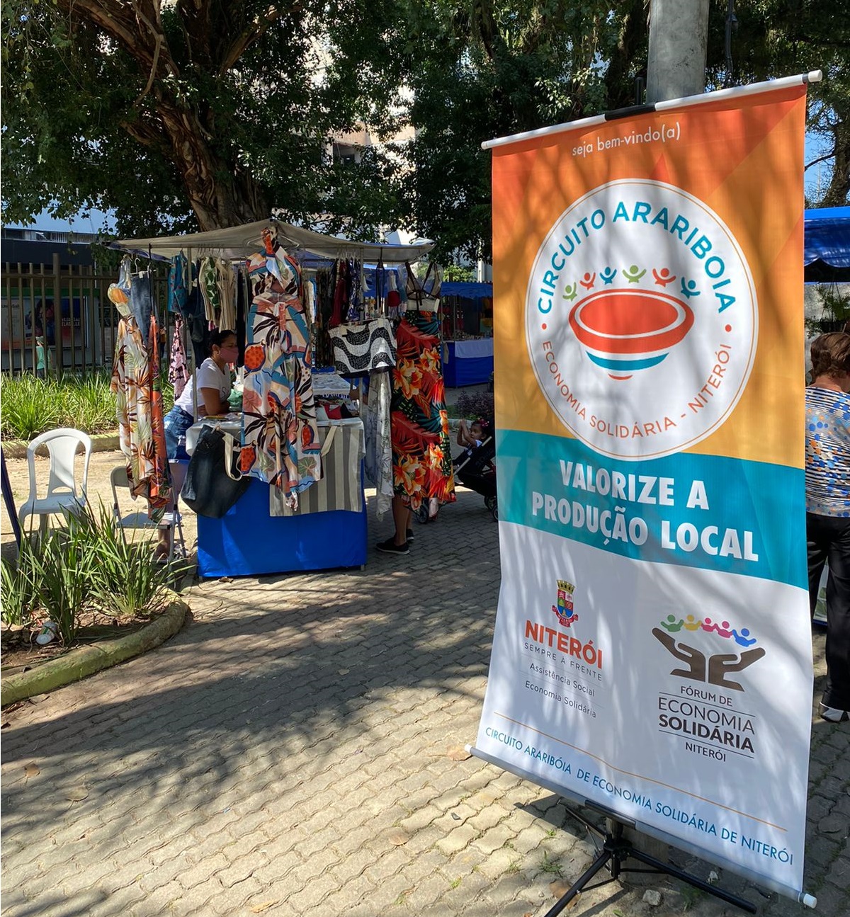 Barraca de roupas estampadas com uma expositora, ao lado de um banner do Circuito Arariboia numa praça arborizada.
