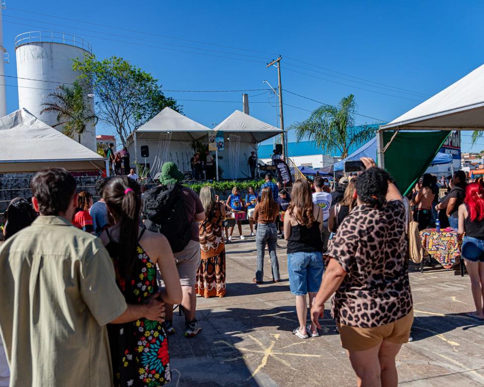 Público de costas assiste a uma apresentação no palco entre tendas brancas, em praça ensolarada.