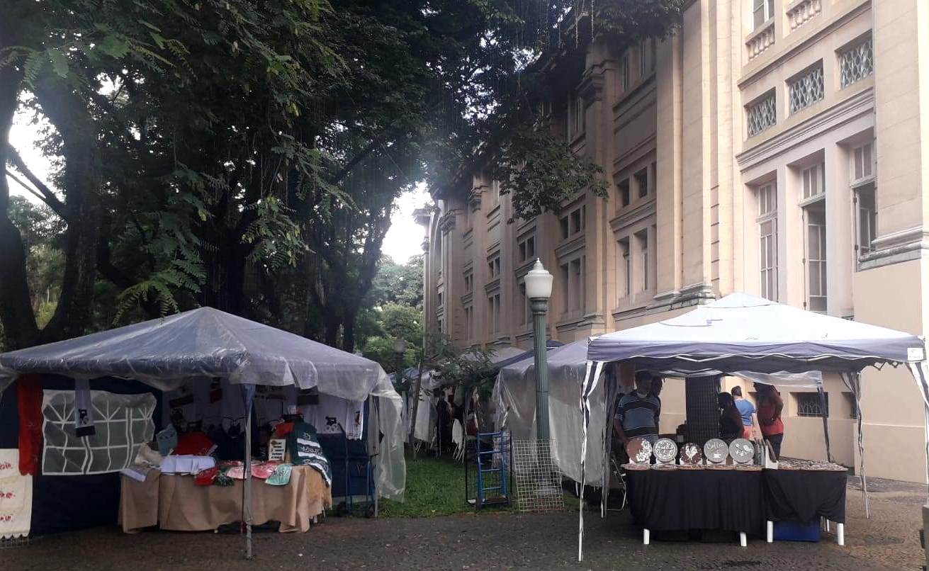 Duas barracas de artesanato sob tendas entre árvores, ao lado de um prédio histórico, com expositores e visitantes ao fundo.