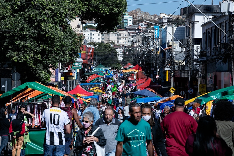 Multidão caminhando pela rua entre barracas coloridas da feira, com morros ao fundo.