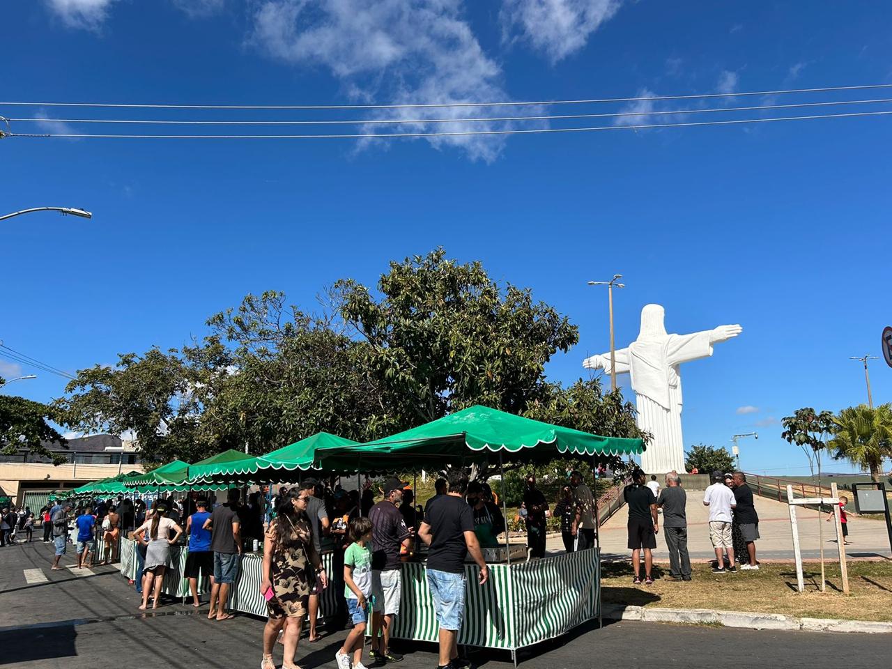 Fileira de barracas verdes e brancas com pessoas circulando na rua, ao lado da estátua branca do Cristo Redentor sob céu azul.