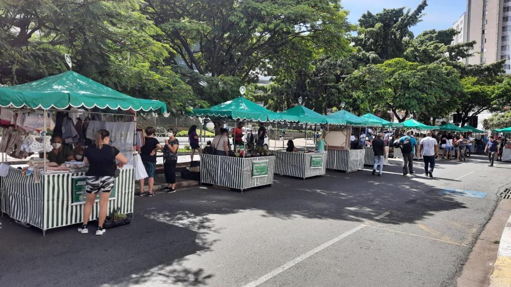 Fileira de barracas com toldos verdes e balcões listrados numa rua, com pessoas olhando as mercadorias e circulando.