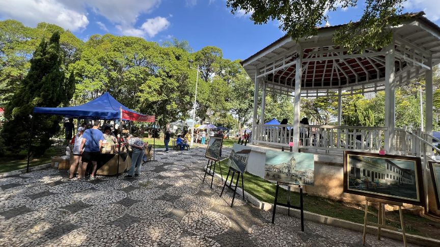 Barraca com tenda azul e quadros em cavaletes ao lado de um coreto numa praça arborizada, com pessoas circulando.