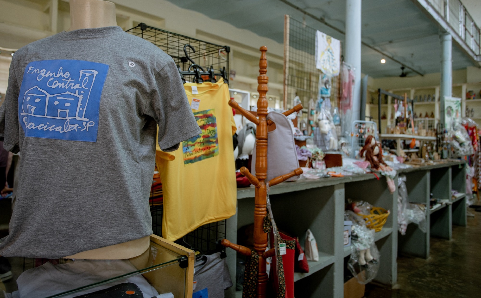 Camiseta cinza com estampa do Engenho Central em primeiro plano e, ao fundo, bancadas com bonecas de pano e outros artesanatos.