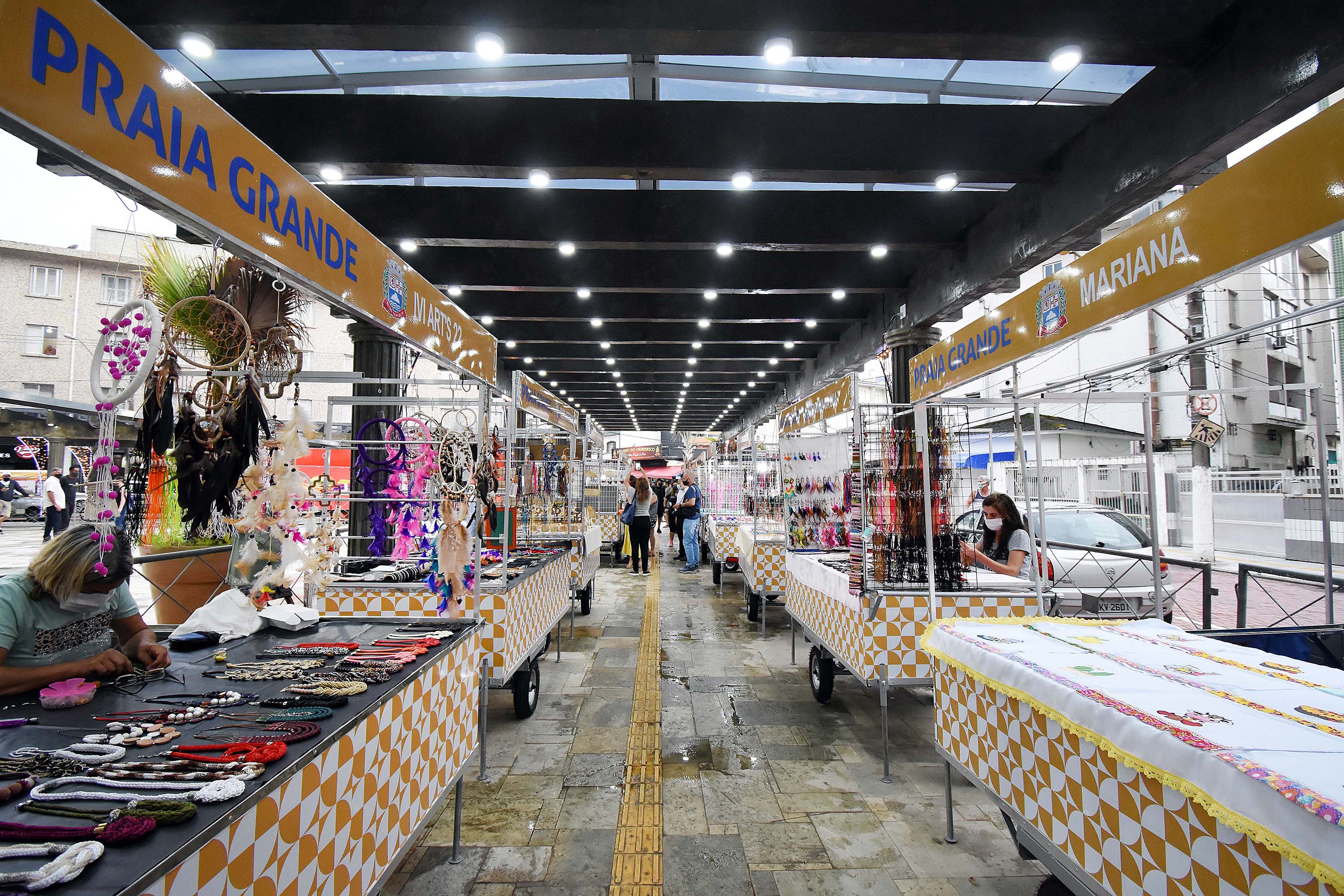 Corredor coberto com barracas de artesanato amarelas e brancas, apanhadores de sonhos e colares expostos.
