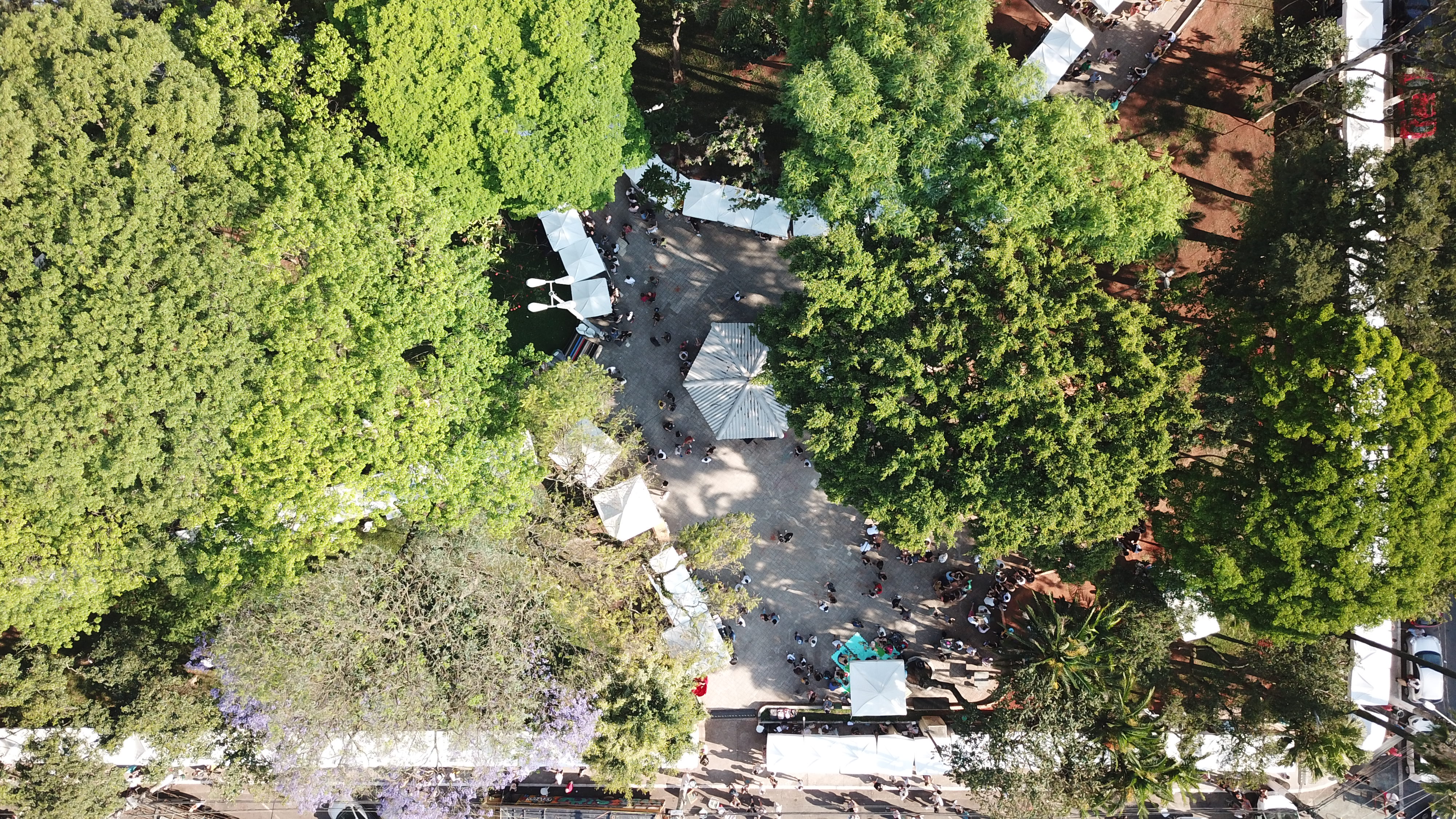 Vista aérea de uma praça arborizada com tendas brancas e pessoas circulando entre as barracas.
