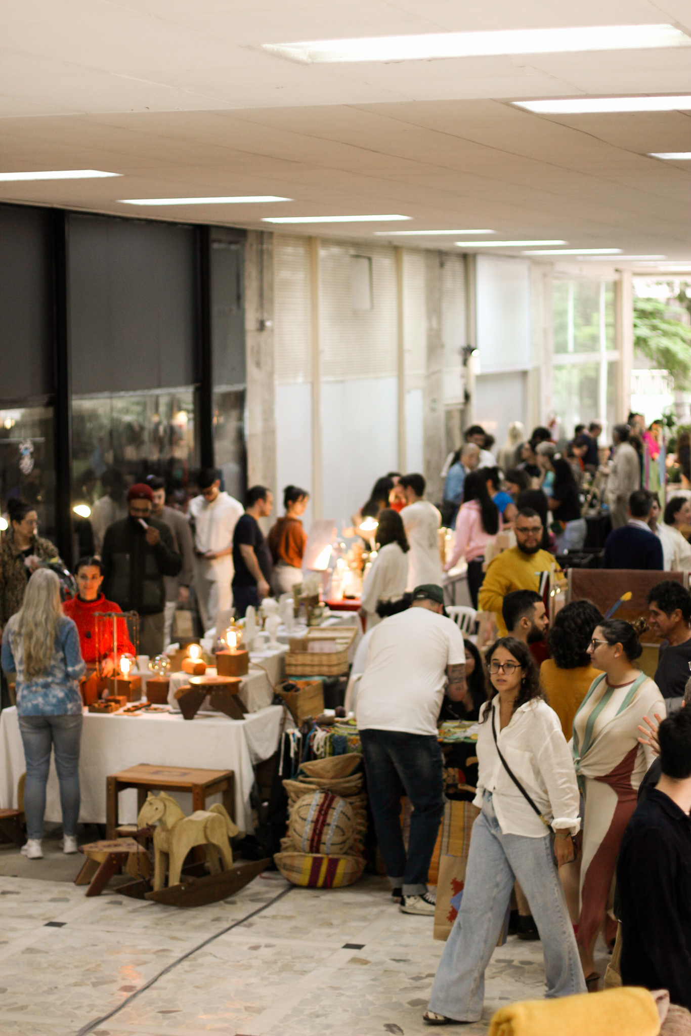 Público circulando entre mesas de artesanato, luminárias e cestos em um salão coberto durante a feira.