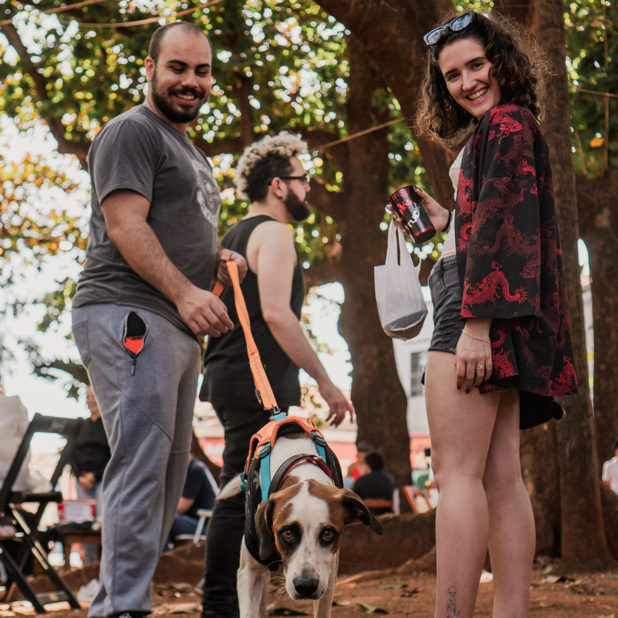 Três pessoas sorrindo com um cachorro de coleira sob árvores da praça, com mesas da feira ao fundo.