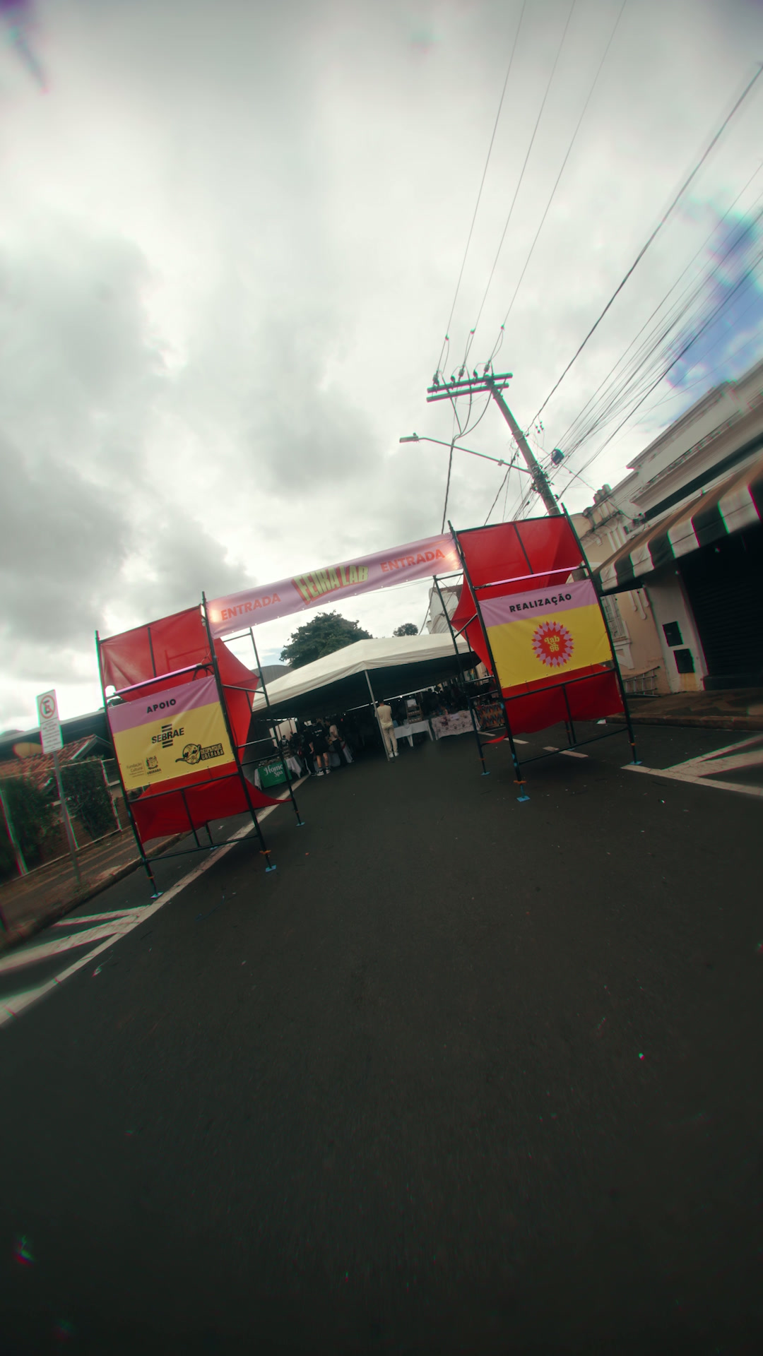 Rua com pórtico vermelho e faixa de entrada da Feira Lab, tenda branca e pessoas circulando ao fundo.