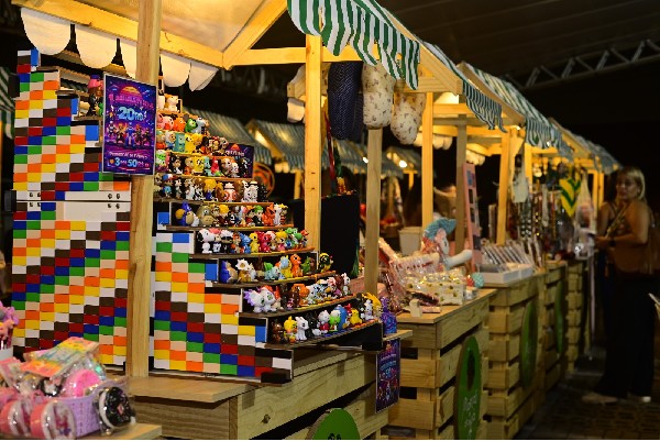 Barracas de madeira com toldos listrados à noite, vendendo bonecos coloridos, com uma pessoa ao fundo.