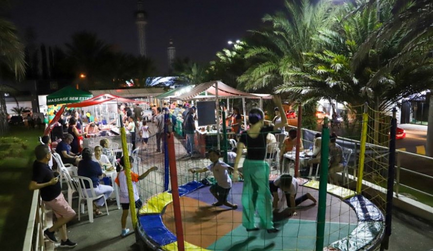 Praça à noite com barracas, mesas com pessoas sentadas e crianças brincando numa cama elástica.