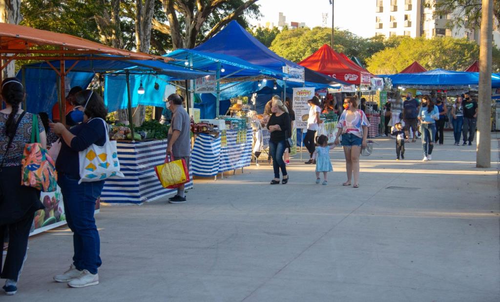 Barracas com toldos azuis e vermelhos vendendo frutas e verduras, com pessoas circulando e comprando na feira ao ar livre.