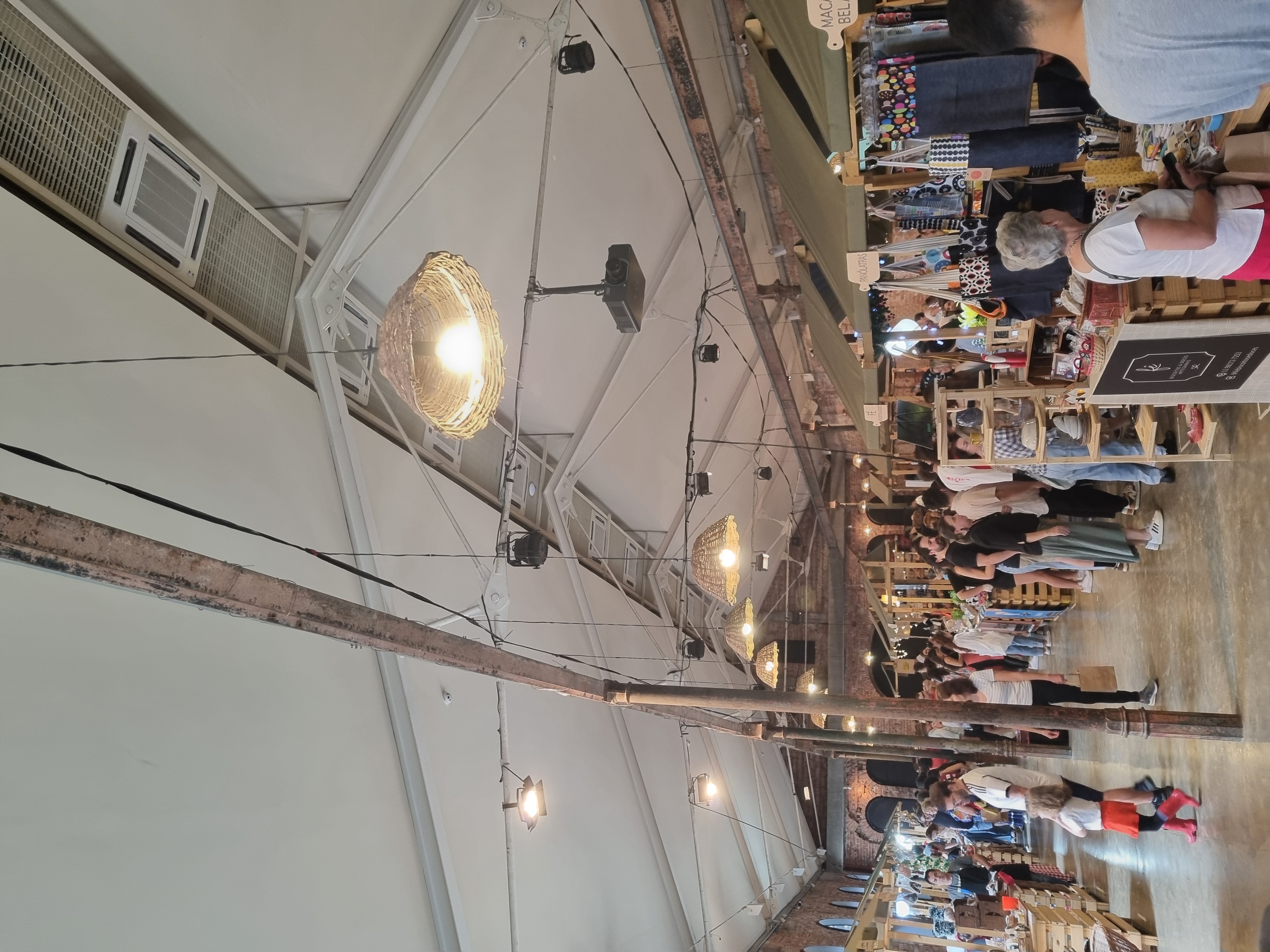 Interior de um galpão de tijolos com barracas de madeira, artesanato pendurado e pessoas circulando sob luminárias de palha.