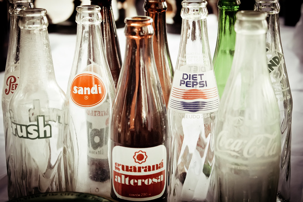 Close de várias garrafas de vidro antigas de refrigerante, entre elas Guaraná Alterosa, Coca-Cola, Crush e Diet Pepsi, alinhadas sobre uma mesa clara.