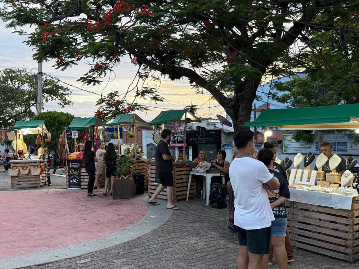 Barracas de toldo verde com artesanato e joias numa praça ao entardecer, com pessoas circulando e comprando.
