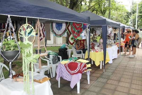 Barracas com tendas azuis na praça, expondo artesanato em crochê e macramê, com visitantes passando.