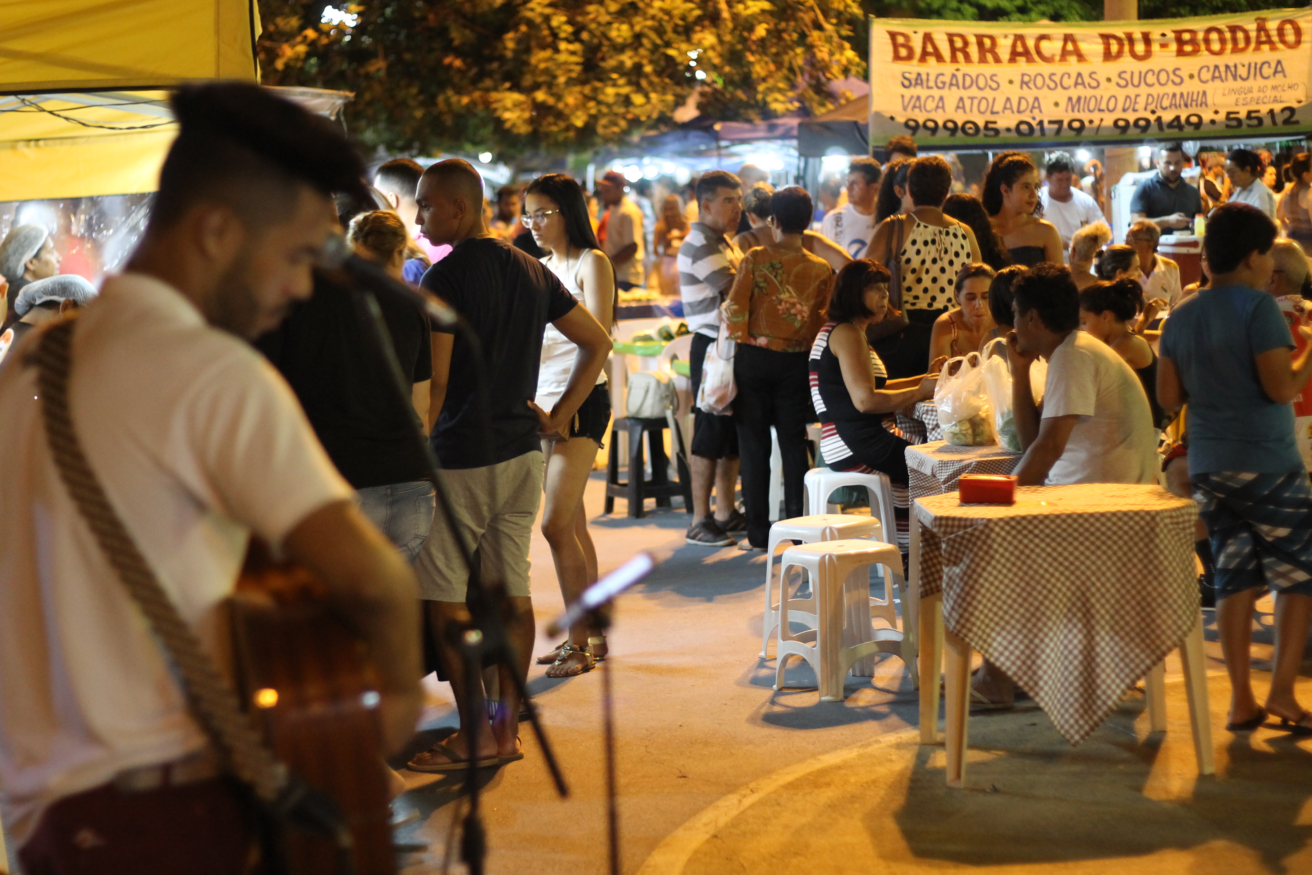 Noite na feira, com público em volta de mesas e barracas e um músico tocando violão em primeiro plano.