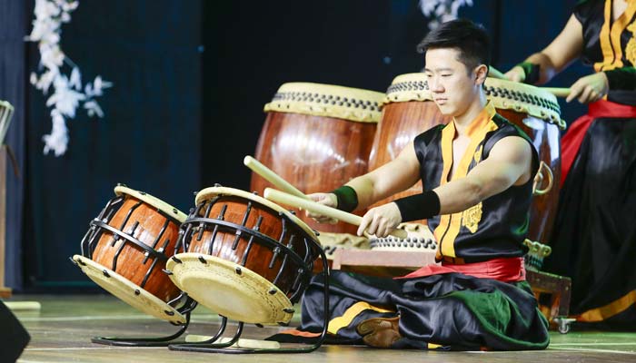 Um tocador de taiko de roupa tradicional preta e amarela toca tambores japoneses no palco.