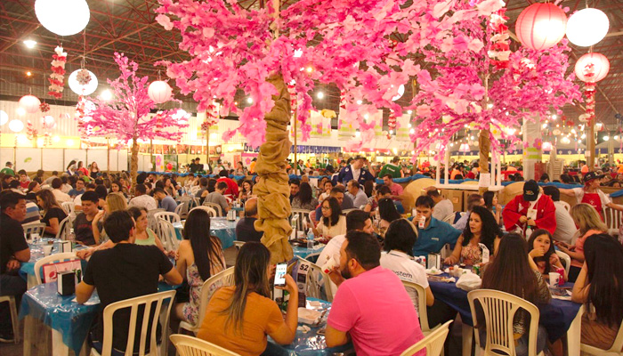Praça de alimentação lotada, com pessoas sentadas em mesas sob cerejeiras artificiais rosas e lanternas japonesas.