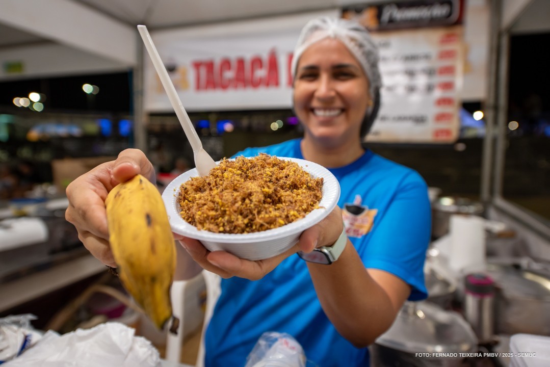 Expositora sorrindo numa barraca à noite, estendendo uma tigela de tacacá com farofa e uma banana.