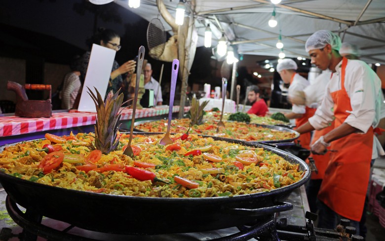 Grandes panelas de paella com legumes em barraca de feira à noite, com cozinheiros de avental laranja atendendo.