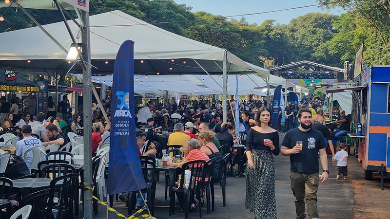 Rua tomada por tendas brancas e mesas com muito público bebendo, com um food truck azul e pessoas caminhando com copos.