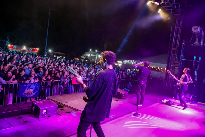 Banda tocando no palco à noite, vista de trás, diante de uma multidão atrás da grade e de tendas iluminadas.