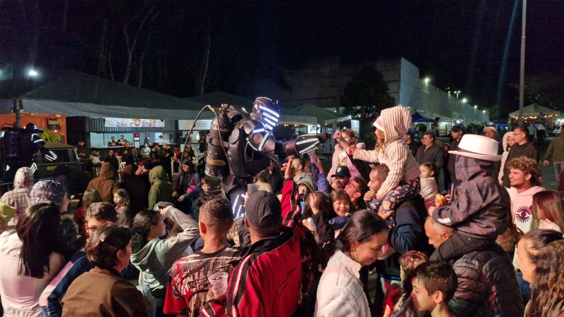 Público reunido à noite diante de um performer com roupa de LED, com tendas iluminadas ao fundo.